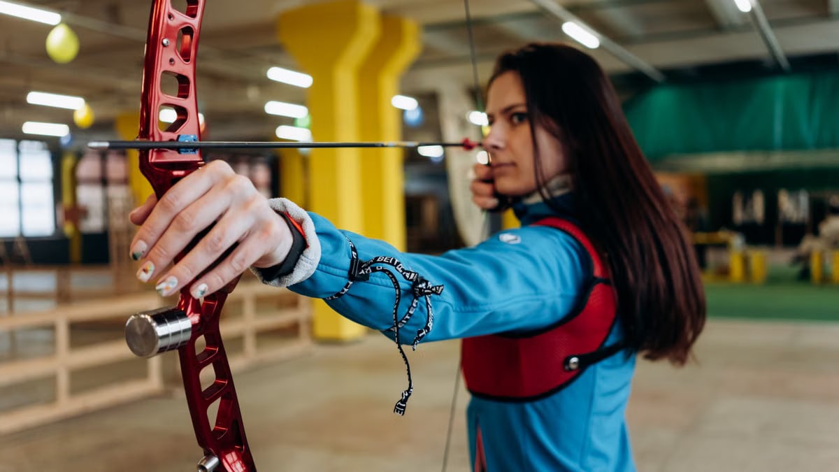 A woman in a blue jacket aims a bow and arrow with concentration during indoor archery practice.